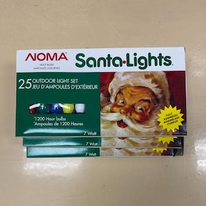 Peut inclure: Une pile de trois boîtes de guirlandes lumineuses d'extérieur NOMA Santa-Lights. Chaque boîte présente une image du Père Noël, le texte 25 Jeu d'ampoules d'extérieur, Ampoules de 1200 Heures et 7 Watt. Une fenêtre montre des ampoules colorées.