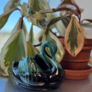 May include: A black and green glazed ceramic swan planter. The swan has a long neck and a detailed design. The planter is sitting on a silver surface.