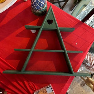 May include: A green, triangular wooden shelf with three horizontal shelves. The top of the shelf has a heart-shaped cutout. The shelf is painted a solid green colour. The shelf is displayed on a red surface.