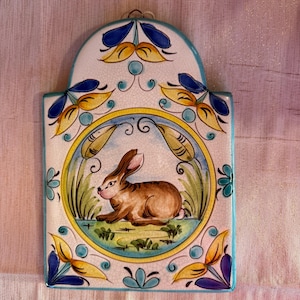 May include: A ceramic wall tile depicting a brown rabbit within a circular frame. The tile has a white base with blue, yellow, and green floral patterns. The tile is rectangular with a curved top and a small hole for hanging.