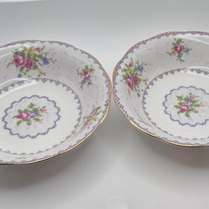 May include: Two white porcelain bowls with a floral design. The bowls have a gold rim and a scalloped edge. The floral design includes pink, blue, and yellow flowers. The bowls are likely for serving or display.