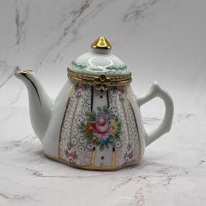 May include: A small white porcelain teapot with a gold lid and floral design. The teapot has a hinged lid and a floral pattern with gold accents.