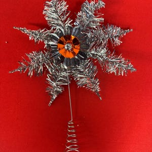 Peut inclure: Un ancien sommet de sapin de Noël avec un motif en étoile. Le sommet présente des branches en guirlande argentée entourant une fleur centrale avec une gemme orange. Un fil d'argent s'étend jusqu'à une base en spirale décorative.