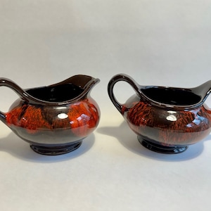 May include: Two brown ceramic cream jugs with red and brown glaze. The jugs have a rounded body and a single handle.