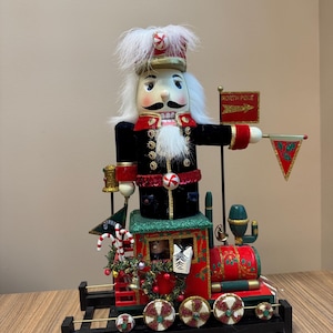 Peut inclure: Un train de Noël décoratif avec un casse-noisette en bois en guise de conducteur. Le train est rouge et vert avec des détails dorés, et les roues sont décorées de paillettes. Le conducteur porte un uniforme noir et un grand chapeau. Le panneau indique "NORTH POLE".