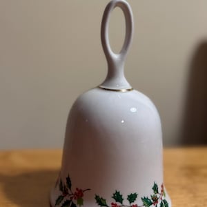 May include: White ceramic bell with a loop handle and a gold rim. The bell is decorated with a garland of green holly leaves and red berries around the base. A festive holiday decoration.