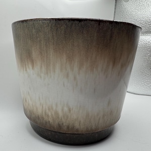 May include: A ceramic plant pot with a gradient design. The top rim is dark brown, transitioning to white in the middle, and ending with a dark brown base. The pot has a cylindrical shape with a slightly flared top.