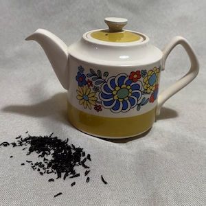 May include: A retro ceramic teapot with a white body, yellow base and lid, and a floral design in blue, yellow, and red. The teapot has a matching lid and handle. A small pile of loose tea leaves is next to the teapot.