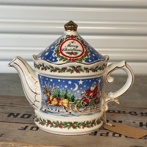 May include: A white ceramic teapot with a lid featuring a gold-tone finial. The body and lid are decorated with a blue night sky, stars, a crescent moon, and a scene of Santa in a sleigh pulled by reindeer. Holly and berry borders frame the design.