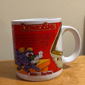 May include: White ceramic mug with a cartoon design. The mug features a purple creature, a computer, and the words "Downtime" and "Bits & Bytes" in a retro style. The mug has a white handle.