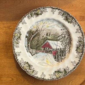May include: A round, off-white decorative plate with a winter scene featuring a red covered bridge, snow, and trees. The rim has a brown and green floral design.
