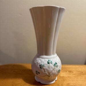 May include: A white porcelain vase with a flared, scalloped rim and a vertical ribbed neck. The rounded base features a textured basket-weave pattern decorated with hand-painted green shamrock motifs and thin brown stems.