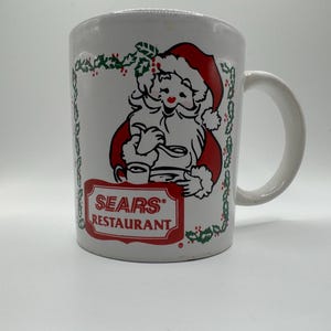 Peut inclure: Tasse en céramique blanche avec une illustration vintage du Père Noël en rouge et noir, tenant un plateau avec des tasses. La tasse porte le texte "SEARS RESTAURANT" dans un cadre rectangulaire rouge. Une bordure verte de houx et de baies encadre l'image du Père Noël.