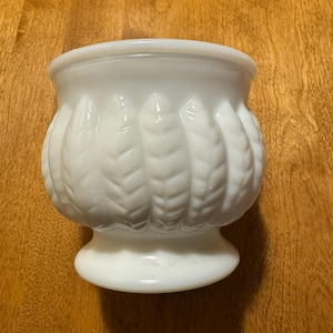 May include: A white ceramic planter with a leaf design. The planter has a wide base and a narrow opening.