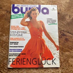 Puede incluir: Un número de la revista Burda de junio de 1992 con una portada que muestra a una persona con un vestido naranja brillante sin mangas con botones dorados. La portada incluye texto como Mode, Handarbeiten, Gastlichkeit y el título Ferienglück.
