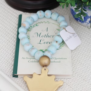 May include: A light blue wooden bead garland with a gold crown charm. The garland is on a book with the text "A Mother's Love" and "An Inspired Celebration of Quotations on Motherhood".