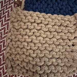 May include: A close-up of a beige knitted pot holder with a dark blue knitted pot holder on top. The pot holders are made of thick rope and have a woven texture.