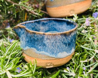 May include: Two handmade ceramic pouring bowls with a blue and tan glaze. The bowls have a small spout and sit on a natural clay base. The bowls are surrounded by green foliage.