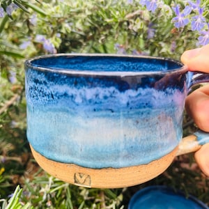 May include: A handmade ceramic mug with a blue and white glaze. The mug has a rounded shape with a handle and a natural, unglazed base. The mug is held in a hand, with greenery and purple flowers in the background.
