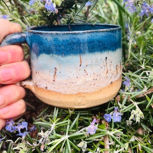 May include: A ceramic mug with a blue and tan glaze. The mug has a rounded base and a handle. The blue glaze transitions from a deep shade at the top to a lighter hue, with tan at the base. The mug is surrounded by green foliage and purple flowers.