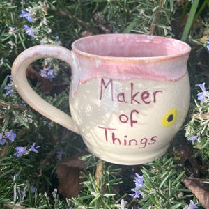 May include: A cream-coloured ceramic mug with a pink rim and handle. The mug has the text "Maker of Things" in dark red lettering, with a small yellow flower. The mug is set against a backdrop of green foliage and small purple flowers.