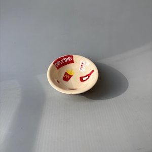 Może przedstawiać: Mała, kremowa ceramiczna miska z ręcznie malowanymi wzorami w stylu fast food. Miska posiada czerwony baner "DRIVE THRU", etykietę "Coke", czerwoną butelkę oraz złote frytki i logo "M". Misa jest okrągła.