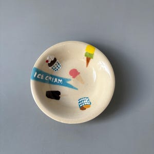Pode incluir: Uma pequena tigela de cerâmica branca com ilustrações coloridas de gelados. A tigela apresenta uma faixa azul com as palavras "ICE CREAM". Os desenhos de gelados incluem cones e picolés em várias cores. A tigela é redonda e tem uma borda ligeiramente elevada.