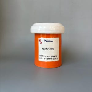 Peut inclure: Une bouteille de pilules en céramique orange avec un couvercle blanc. L'étiquette indique "KLONOPIN" et "WHEN A DEEP BREATH JUST DOESN'T CUT IT." La bouteille porte également le symbole de la pharmacie et le mot "Pharmacy".