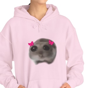 Può includere: Una felpa con cappuccio rosa chiaro con tasca frontale e cappuccio con coulisse. La felpa presenta una grafica in stile acquerello di una creatura triste con fiocchi rosa. La modella indossa pantaloni neri.