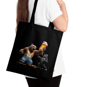 Puede incluir: Bolso tote negro con una ilustración de dibujos animados de un oso y un gorila. El oso sostiene una pinta de cerveza, brindando con el gorila. El oso lleva una camiseta sin mangas y un peto. El bolso tiene asas negras.