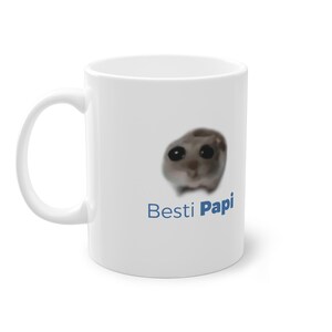 Könnte beinhalten: Weiße Keramiktasse mit einem verschwommenen Bild eines kleinen Tieres mit großen Augen und dem Text "Besti Papi" in Blau.