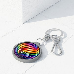 Könnte beinhalten: Ein grauer Schlüsselanhänger mit einem runden Anhänger, der ein lebendiges Regenbogen-Wirbel-Design und den Text "LGBTQ+" zeigt. Der Schlüsselanhänger hat einen silbernen Ring und Karabiner.
