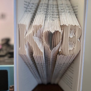 May include: A book with the letters "K" and "E" folded into the pages, with a heart shape in the middle. The book is open and standing upright on a wooden surface.