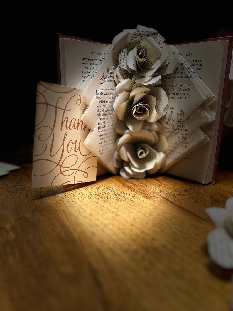 May include: An open book with pages folded into a decorative design, adorned with three paper roses. A card with the words "Thank You" in elegant script is placed in front of the book, all on a wooden surface.