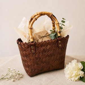 Puede incluir: Una cesta tejida marrón con asa de bambú, llena de papel de seda y un regalo envuelto. La cesta tiene forma rectangular y está decorada con una ramita de vegetación. Una flor blanca y pequeñas flores blancas están en primer plano.