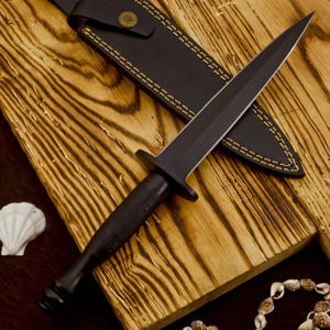 Op de afbeelding: Een zwarte dolk met een dubbelzijdig lemmet en een getextureerde handgreep, rustend op een houten oppervlak. De dolk wordt vergezeld door een zwarte leren schede met gele stiksels. Een zeeschelp en een schelpenketting zijn ook zichtbaar.