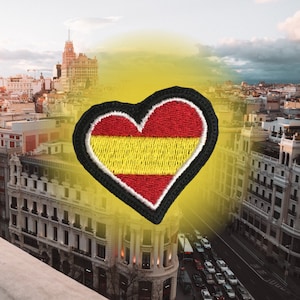 Puede incluir: Un parche en forma de corazón rojo y amarillo con un contorno negro, que representa la bandera española, se superpone sobre una imagen borrosa de un horizonte urbano.