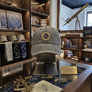 May include: A gray baseball cap with an embroidered eclipse design. The cap features a dark circle with a gold border and a sun-like detail. The cap is displayed on a metal stand in a shop setting.