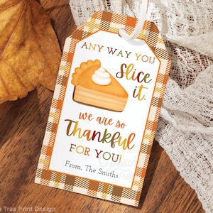 May include: A gift tag with a pumpkin pie illustration and the text "Any way you slice it, we are so thankful for you!" The tag has a plaid pattern in orange and brown. The tag is attached to a white string and is on a wooden surface.