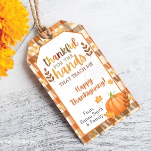 Op de afbeelding: Thanksgiving cadeaulabel met een oranje en bruin ruitpatroon. Het label leest "dankbaar voor de handen die me leren Happy Thanksgiving!" met een pompoen- en bladerillustratie. Een boeket oranje bloemen staat op de achtergrond.