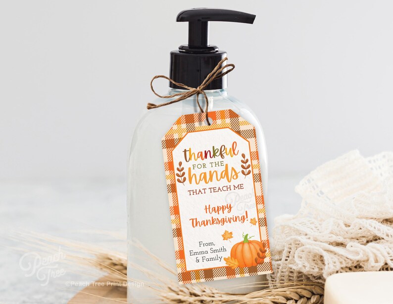 Op de afbeelding: Een doorzichtige vloeibare zeepdispenser met een zwarte pomp en een decoratief label. Het label heeft een ruitpatroon in oranje en wit, met de tekst "thankful for the hands that teach me Happy Thanksgiving!" en een pompoenillustratie.