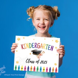 May include: A smiling child holds a white sign with the word "Kindergarten" in rainbow colors. The sign features a graduation cap, diploma, and the text "Class of 2025", with colorful crayons at the bottom and star accents.
