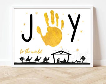 May include: A white printable Christmas card with the word "JOY" in black letters. A gold handprint is in the center of the card. The text "to the world" is below the handprint. A black silhouette of a stable with three wise men and a donkey is at the bottom of the card.