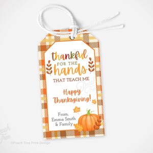 Op de afbeelding: Thanksgiving cadeaulabel met een oranje en bruine geruite rand. Het label leest "dankbaar voor de handen die me leren" en "Fijne Thanksgiving!" met een pompoen- en bladerillustratie. Inclusief een wit touwtje.