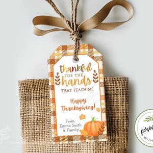 Op de afbeelding: Thanksgiving cadeaulabel met een geruite rand en de tekst "dankbaar voor de handen die me leren". Een pompoenillustratie en de woorden "Happy Thanksgiving" zijn ook te zien. Het label is met een bruin lint aan een jute zak gebonden.
