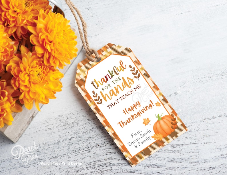 Op de afbeelding: Thanksgiving cadeaulabel met een oranje en bruin ruitpatroon. Het label leest "dankbaar voor de handen die me leren Happy Thanksgiving!" met een pompoen- en bladerillustratie. Een boeket oranje bloemen staat op de achtergrond.