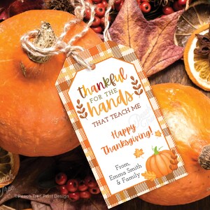 Op de afbeelding: Twee oranje pompoenen met een Thanksgiving-cadeaulabel. Het label luidt "dankbaar voor de handen die me leren Vrolijk Thanksgiving!" met een pompoenillustratie en herfstbladeren. Gedroogde sinaasappelschijfjes en bessen zijn ook zichtbaar.