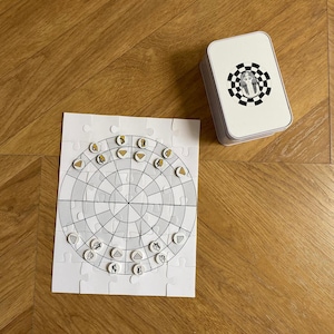 May include: A white puzzle board with a circular grid and 24 small, round, white tiles with gold accents. The tiles are arranged in a circle around the grid. A white box with a black and white image of an Egyptian pharaoh is in the upper right corner of the image.