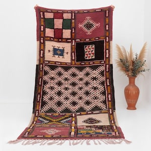 May include: A rectangular rug with a patchwork design featuring geometric patterns in pink, burgundy, black, and cream. The rug has a fringed edge and is displayed against a white background. The rug's dimensions are not visible.