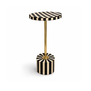 Puede incluir: Una mesa auxiliar con un tablero circular y una base cilíndrica, ambos decorados con un patrón de rayas negras y crema. Una varilla metálica delgada de tono dorado conecta el tablero con la base, creando una estructura vertical.